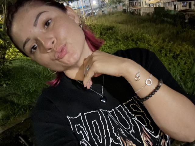 KatherineToro with pink-dyed hair, pouty lips, black shirt, necklace, bracelet, and earrings, making a kissy face, holding her chin. Nighttime, grassy background.