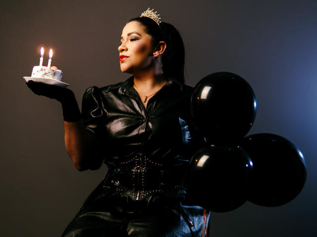 Molly Reed con un conjunto de cuero negro, una corona, guantes negros, sosteniendo un pastel con velas, globos negros, y una mirada sexy y segura. Se ven los senos debajo de la camisa de cuero, fondo oscuro..
