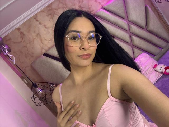 IsaDoux, a Latina woman with long black hair, wears clear glasses and a pink satin camisole with thin straps, revealing a small cleavage. She touches her chest with her right hand, lips slightly parted with a subtle smile. Her skin is light brown. The pink satin contrasts with her dark hair and clear glasses. The image is brightly lit with pink and purple lighting, creating a soft, intimate atmosphere. Her nails are painted pink.