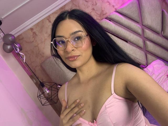 IsaDoux with long black hair, pink glasses, and pink bra, covering her small tits with her hand, in a pink-lit bedroom. Her cute face has a slight smile.