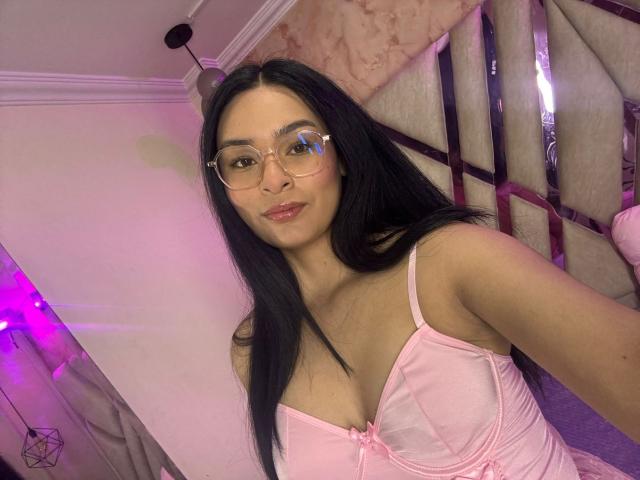 A photo of IsaDoux with long black hair and clear glasses. She wears a pink satin bra with a small bow in the center. Her skin is light brown, and she has pink lipstick. IsaDoux's bra highlights her small to medium-sized breasts. She's taking a selfie in a room with pink and purple lighting. The background includes a bed with beige and pink patterned pillows and a geometric shelf. The image is clear and focused on IsaDoux's upper body.