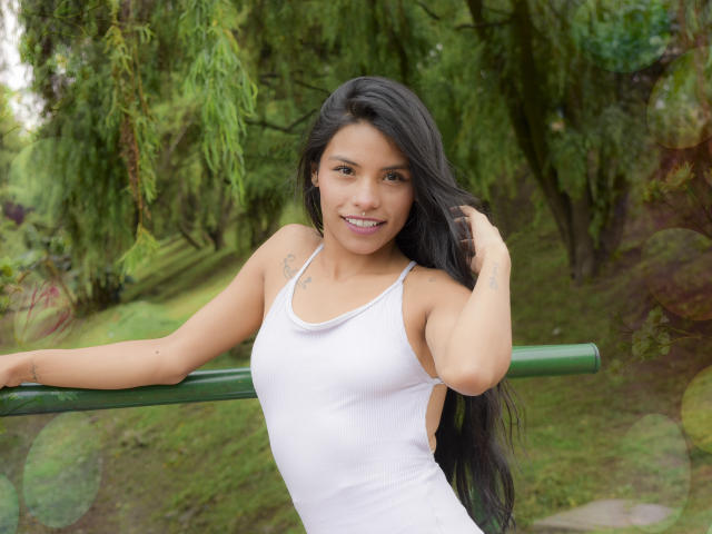 ClarisCampbell with long black hair, smiling, wearing a white tank top, leaning on a green pole, her ass slightly visible, in a green, leafy park.