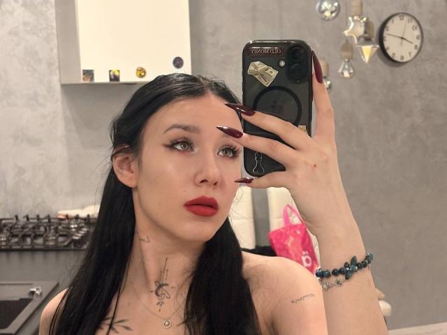 NikkiGoldX takes a mirror selfie in a modern kitchen. She has long black hair, red lipstick, and dark red nails. Her light skin contrasts with her black hair. She holds a black phone with a sticker on the back in her right hand, which also has a blue bracelet. She has tattoos on her neck and chest. The background includes a white cabinet, a clock, and hanging ornaments. Her expression is neutral. The photo is a selfie.