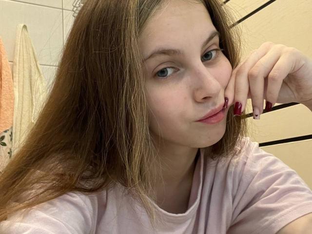 IoneTrosper has long brown hair, pale skin, blue eyes, and pink lipstick, resting her chin on her hand with red nails, wearing a pink shirt.