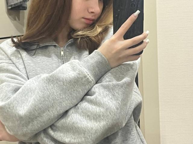IoneTrosper with light brown hair, wearing a grey zip-up hoodie, taking a selfie with a black phone, showing a white ring on her finger, crossed arms, subtle pout.