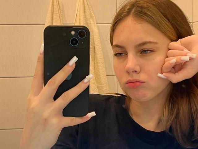 IoneTrosper with puffy lips, holding a black phone, wearing a black shirt, and long white nails, looking into the camera with a slightly annoyed expression.