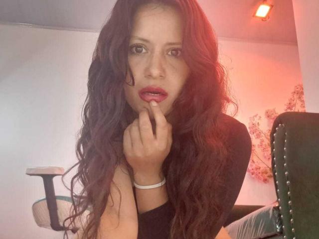 AriannaSeete with long, wavy brown hair and parted lips, looks directly at the camera with a slightly open mouth. She has her right hand near her chin, with fingers gently touching her lips. She wears a black long-sleeve top and a white wristband. The light has a redish hue, creating a warm glow on her face. The background is blurred, focusing on AriannaSeete's expression and hair texture. The image has a casual, candid feel.