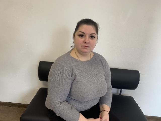 AdelinaMaster, a chubby woman with dark hair in a ponytail, wearing a gray long-sleeve shirt, sits on a black chair, looking serious with her hands on her lap.