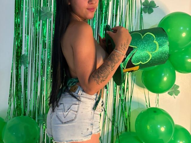 EimmyyRousee with long black hair, topless, showing her tits, wearing white denim shorts, holding a green glittery St. Patrick's Day roll, surrounded by green balloons and green streamers.