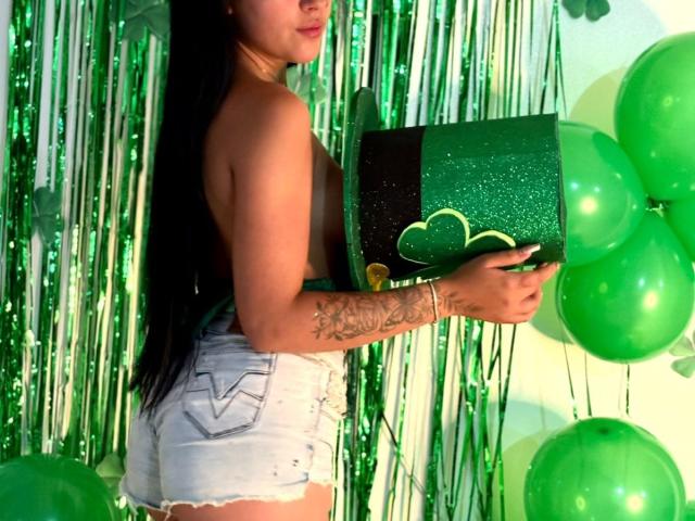 EimmyyRousee with long black hair, topless, holding green glitter box, wearing frayed white shorts, green balloons, green streamers, and a tattoo on her right arm.