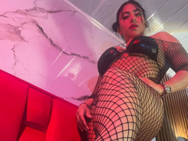 DiannaRosse in black fishnet dress, shiny black bra, and red lighting, standing with one leg raised, showing her curvy ass and large tits, wearing a watch.
