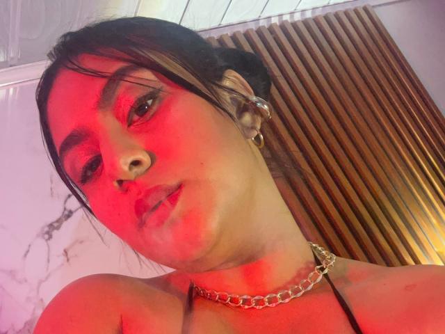 DiannaRosse has red-lit skin, black hair in a messy bun, and a nose ring. She's wearing a gold chain necklace, looking at the camera with a slight smile.