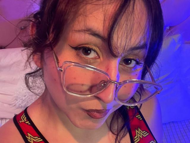 MaddieLesYoux with glasses, brown eyes, and dark hair, wearing a red and black top with yellow designs, looking into the camera with a slight pout.