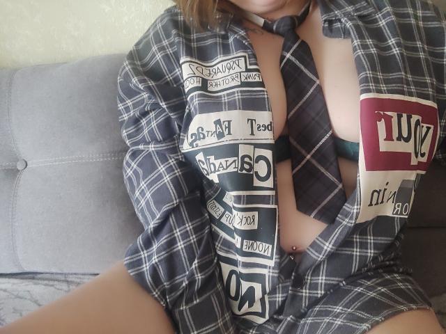 MinistraX wearing an unbuttoned plaid shirt, exposing her tits and a black tie with white text, sitting with her legs apart on a gray couch.