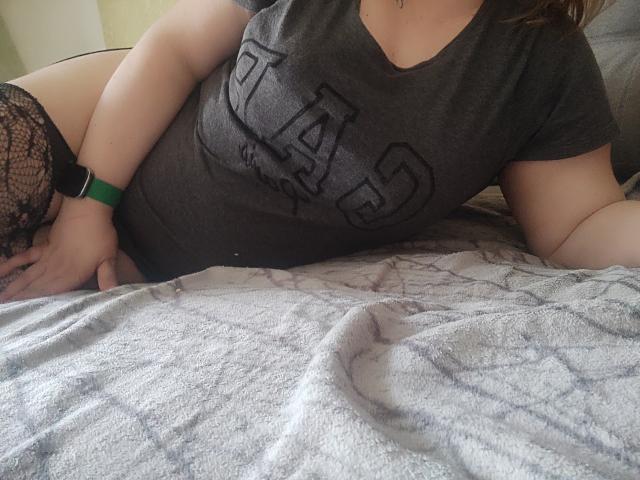 MinistraX lies on a bed, wearing a gray "LAS" t-shirt, black lace stockings, and a green wristband. Her hand is on her chubby ass, and her tits are visible.