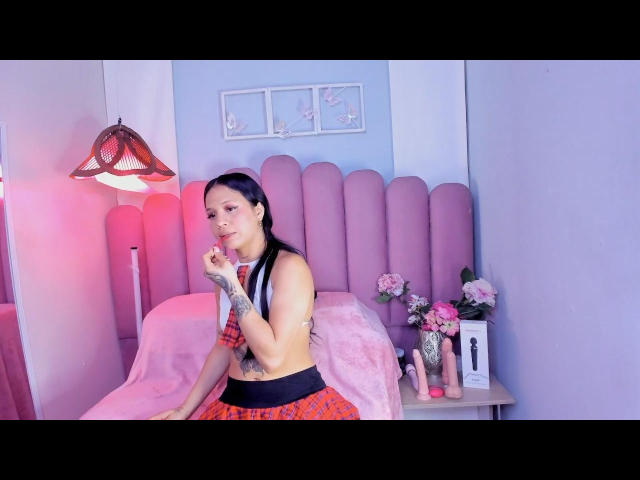 LuciannaVega with black hair, small tits, tattoo on left arm, wearing red plaid skirt and black top, finger in mouth, sitting on pink bed, pink lamp on left.