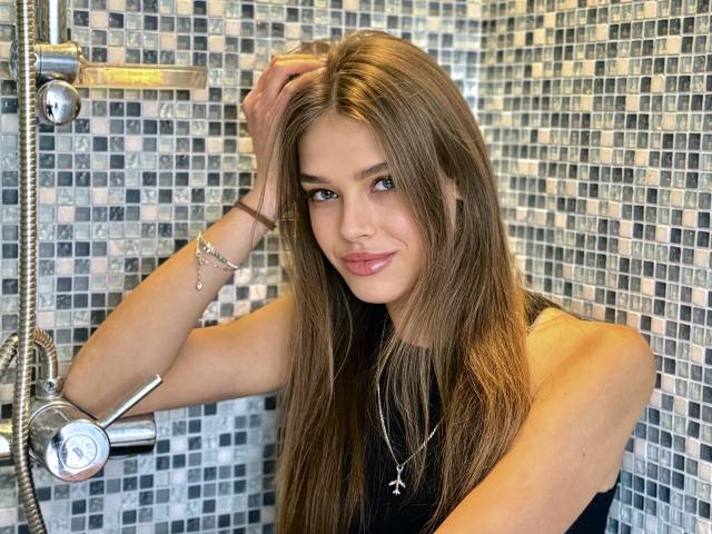 MelissaCrayzy has long, straight brown hair, and she's standing in a shower with a mosaic tile wall. She's wearing a black sleeveless top and a silver necklace with a star pendant. Her right hand is in her hair, and she's smiling with slightly parted lips. She has light skin, and her brown eyes are focused on the camera. A silver showerhead is on the left. She's wearing a thin brown bracelet on her right wrist. The image has a clear, detailed quality.