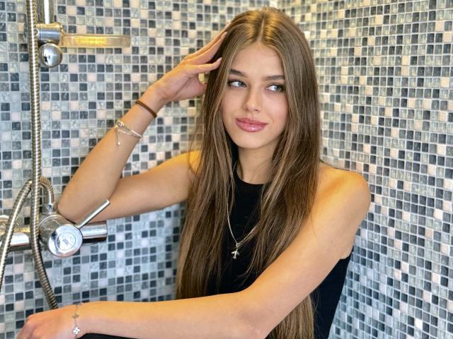 MelissaCrayzy is in a shower with a mosaic tile background. She has long, straight brown hair, light skin, and is wearing a black sleeveless top. She is looking to the side with a slight smile, touching her hair with her right hand. She wears a silver necklace with a star pendant and a black bracelet on her right wrist. The shower has a silver showerhead and a handheld shower attachment. Her eyes are light blue. The image is clear and well-lit.
