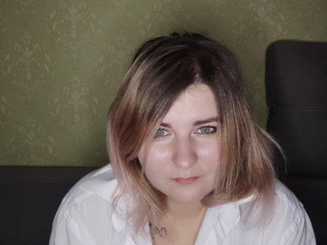 MinistraX has pale skin, green eyes, and shoulder-length brown hair with blonde highlights. She's wearing a white shirt, looking slightly up with a subtle smile. Boobs not visible.