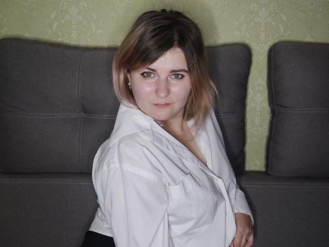 MinistraX with medium breasts, white shirt, black pants, and brown hair, sitting on gray couch, looking directly at camera with slight smile. Green patterned wall in background.