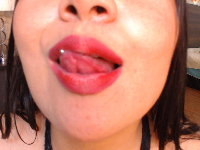 HannahMolly's face with her tongue out, lips slightly parted, showing a small silver tongue piercing, and glossy red lipstick. Her black hair is partially visible.