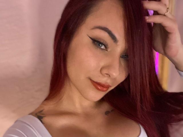 Close-up of ValeryHarllow with straight, dark red hair, slightly parted to the side. She has light skin, thick black eyebrows, and is wearing black eyeliner with a sharp wing. Her lips are glossy with a reddish-pink color. She's looking to the right with a subtle smile. Her right hand is raised, fingers gently touching her hair. She has tattoos on her right shoulder and left collarbone. She's wearing a white top with a low neckline. The background is a soft pink, slightly out of focus. The image has a warm, slightly blurred texture.