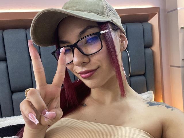 ValeryHarllow, a young woman with light brown skin and red-dyed hair, wears a green cap and black glasses. She makes a peace sign with her right hand, showing pink, glittery nails. She has a small tattoo on her right shoulder and wears large, silver hoop earrings. Her pink lips are slightly parted. The composition focuses on her face and upper torso. The image is clear with a soft, warm light.