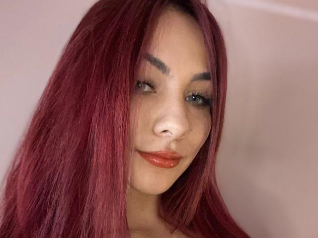 Close-up of ValeryHarllow with straight, vibrant red hair covering her right eye. She has light skin, subtle freckles, and dark, arched eyebrows. ValeryHarllow wears glossy, orange-red lipstick and has long, thick eyelashes. Her expression is neutral with a slight, confident smile. The image focuses on her face, with her hair slightly parted to reveal her left eye. The lighting is soft, highlighting her facial features. The background is blurred, keeping the attention on ValeryHarllow's face.