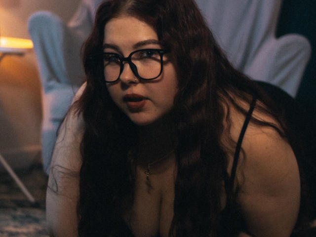 EffyRain has long, wavy brown hair, black glasses, and is wearing a black dress with a low neckline, exposing her big tits. She's on her knees, looking forward.