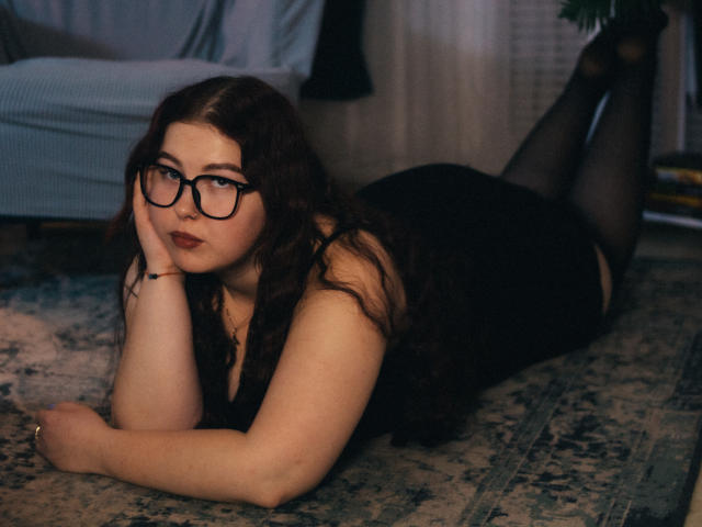 EffyRain is lying on a rug, wearing black glasses, black lingerie, and sheer stockings, with her ass up and face resting on her hand, looking seductive.