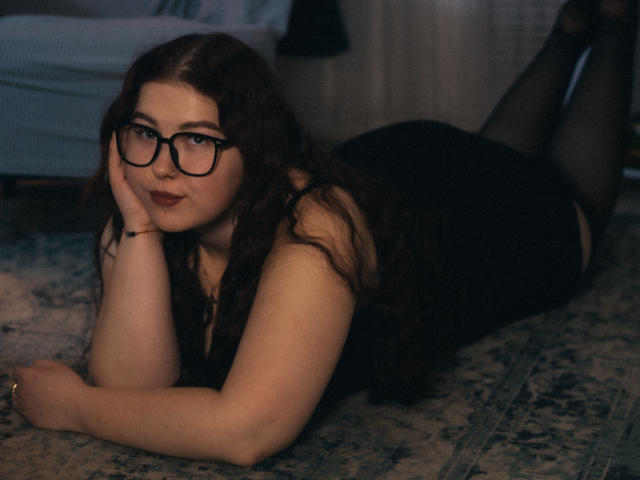 EffyRain with glasses, black dress, and thigh-highs, lying on a rug, butt up, looking at camera, hand under chin, dark red hair, fair skin, subtle smile.
