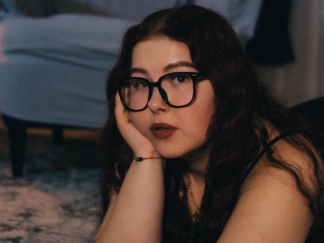 EffyRain with wavy brown hair, wearing black glasses, resting her chin on her hand, looking slightly bored, with red lipstick and a black bracelet on a patterned rug.