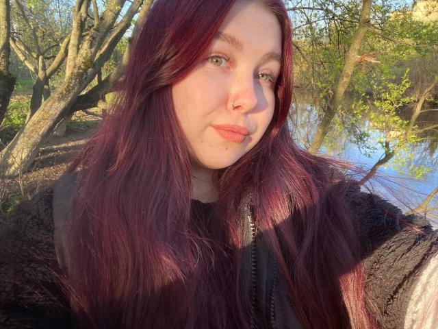 EffyRain with long, red-brown hair, green eyes, nose ring, and light skin, wearing a dark jacket, standing outdoors with sunlight on her face, and a tree in the background.
