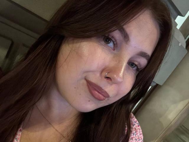 EffyRain has brown hair, blue eyes, a nose ring, and pinkish lipstick with small freckles on her cheek. Her pink floral top is visible. She's smiling slightly.