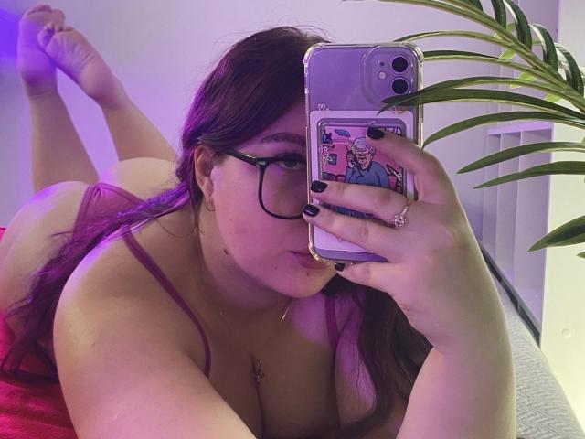 EffyRain, lying on her stomach, wearing purple lingerie, taking a selfie with her phone, showing her tits, butt up, purple hair, glasses, black nails, and a plant in the background.