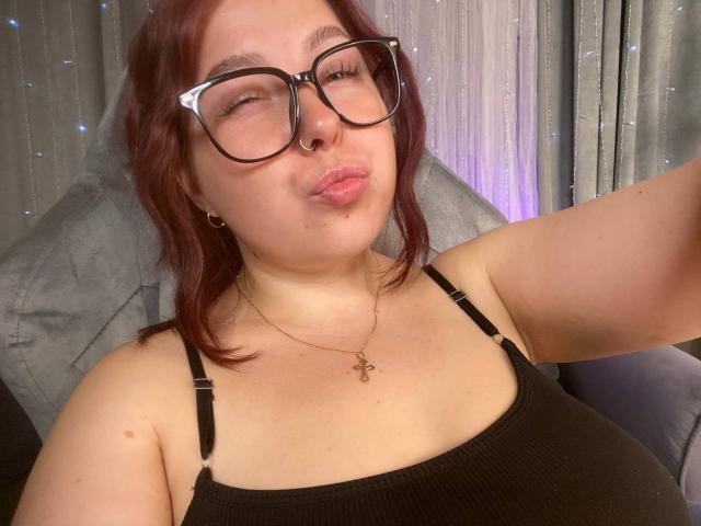 EffyRain with puffy lips, black glasses, red hair, nose ring, black tank top, showing her tits, gold necklace, and a cute expression, taking a selfie.
