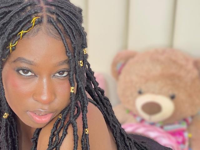 KhaliMaxwell with dark brown skin, black dreadlocks, yellow hairpins, and a serious expression. Her small tits are visible, and a brown teddy bear is in the blurry background.