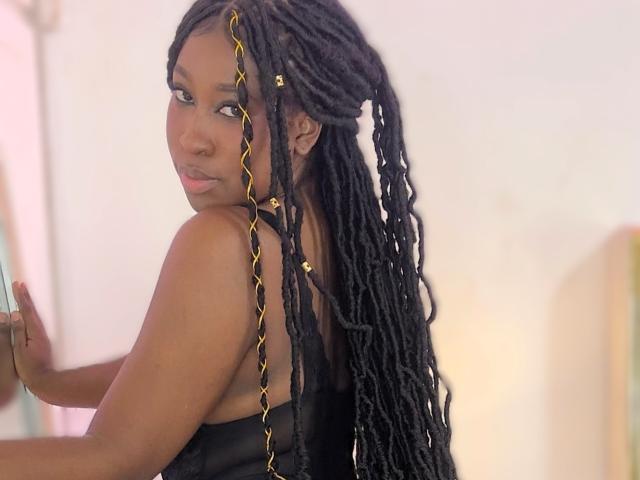 KhaliMaxwell with long black twisted braids, gold extensions, and a black tank top, looking over her shoulder, showing her ass. Soft pink light.