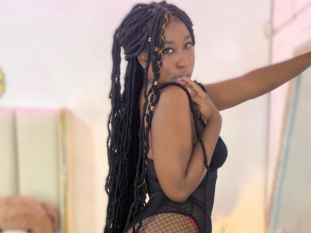 KhaliMaxwell with long black braids, wearing a black fishnet top, looking over her shoulder, hand near her mouth, showing her curvy ass and small, perky tits.