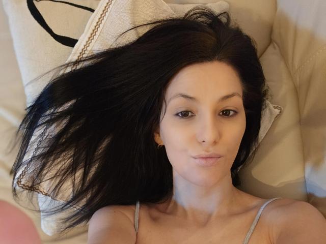 JoliDanielle acostada sobre un cojín, con el cabello negro extendido, ojos marrones, nariz pequeña, vistiendo una camiseta gris, una sonrisa sutil, pequeños aretes dorados, una luz suave en su rostro..