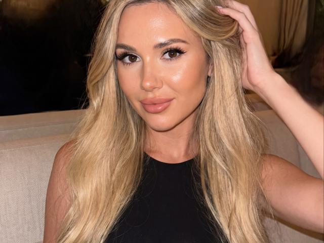 Close-up of NensySafrons, a blonde woman with long, wavy hair, lightly touching her hair with her right hand. She has tan skin, full lips with nude lipstick, and prominent, dark, arched eyebrows. Her brown eyes are accentuated with thick, black eyeliner and long, dark eyelashes. She wears a black, sleeveless top. The lighting is warm, highlighting her smooth, shiny skin. The image has a slightly blurred background.