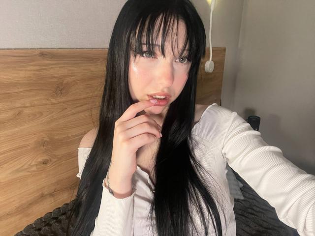 AmaiRin with long black hair, white off-shoulder top, and pink lipstick, biting her finger with a cute, sexy expression, showing off her small, cute tits. Bedroom selfie.