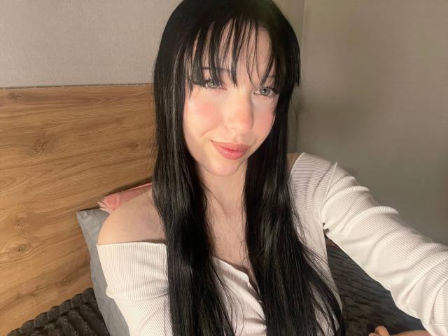 AmaiRin with long black hair, white off-shoulder top, smiling at camera, showing her tits, pink lipstick, pale skin, and blue eyes, sitting on bed with wooden headboard.