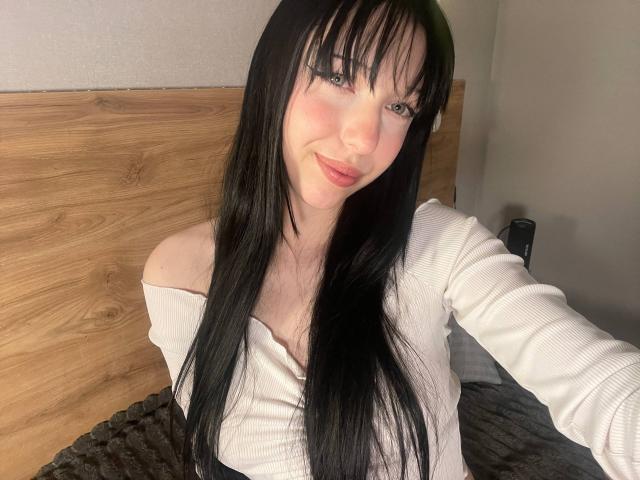 AmaiRin with long black hair, off-shoulder white top, and pink lipstick, smiling at the camera, showing her small tits, sitting on a bed with a wooden headboard.