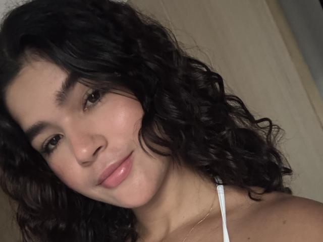 ScarlletZhen has curly dark brown hair, light skin, and pinkish lips, wearing a white strap, looking softly at the camera with a slight smile.