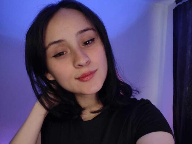 AnnieLamberth is a young woman with light skin and black, shoulder-length hair. She has a slight, confident smile with pink lips. Her eyebrows are thin and arched, and she has subtle winged eyeliner. She wears a simple black T-shirt. Her right hand is raised to her head, touching her hair. The image has a blue-purple gradient in the background, with a dark curtain on the right. AnnieLamberth's face is centered, and she looks directly at the camera. The overall composition is casual and intimate.