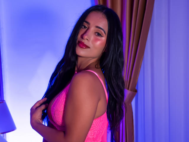 NicolSantos with long black hair, pink lace bra, and red lipstick, holding her right boob, smiling, blue and purple lighting, golden curtains in the background.