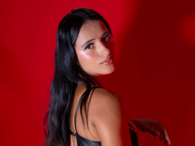 NicolSantos with long black hair, shiny pink lips, and subtle makeup, wearing a black strap top, looking over her shoulder with a sexy, confident smile. Red background.