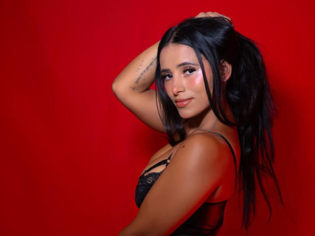 NicolSantos has long black hair, partially held up with her right hand. She wears a black lace bra, highlighting her medium-sized breasts. Her skin is tan, and she has a small tattoo on her left forearm. She looks over her shoulder with a slight smile, eyes partially closed. The red background is solid and contrasts with her hair and skin. Her makeup includes pink eyeshadow and glossy lips. The lighting is even, emphasizing her facial features and hair texture. She has a confident, sexy look.