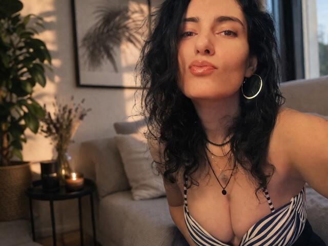 AddictedToUTS avec ses cheveux noirs bouclés, ses boucles d'oreilles, et son haut rayé qui met en valeur sa poitrine généreuse, ses lèvres pulpeuses et un léger sourire, assise dans une pièce confortable avec des bougies..